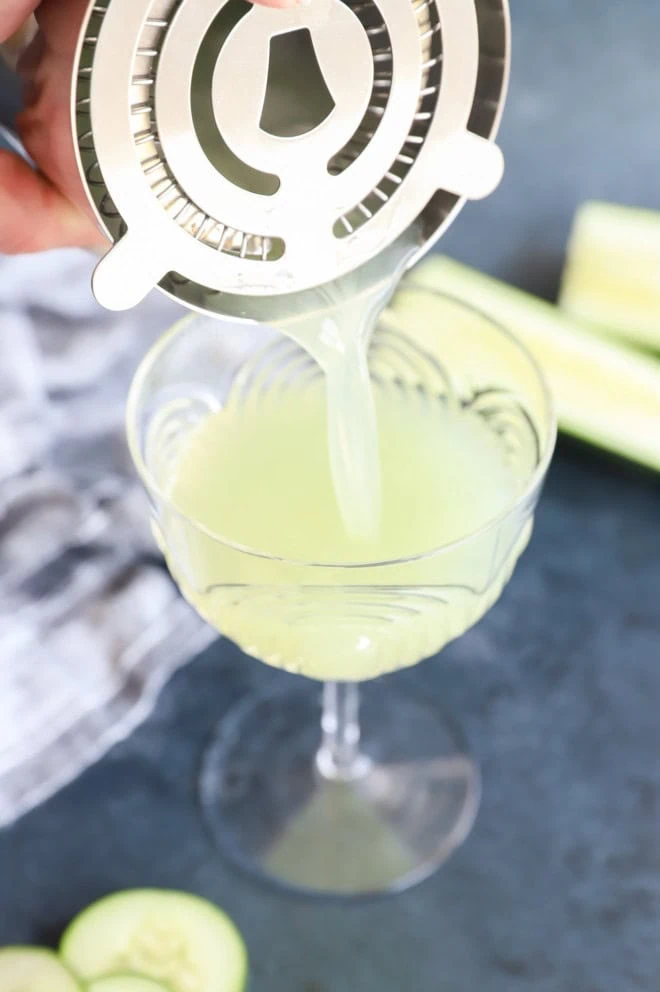 pouring a cucumber martini into a couple glass with cocktail shaker and cocktail strainer