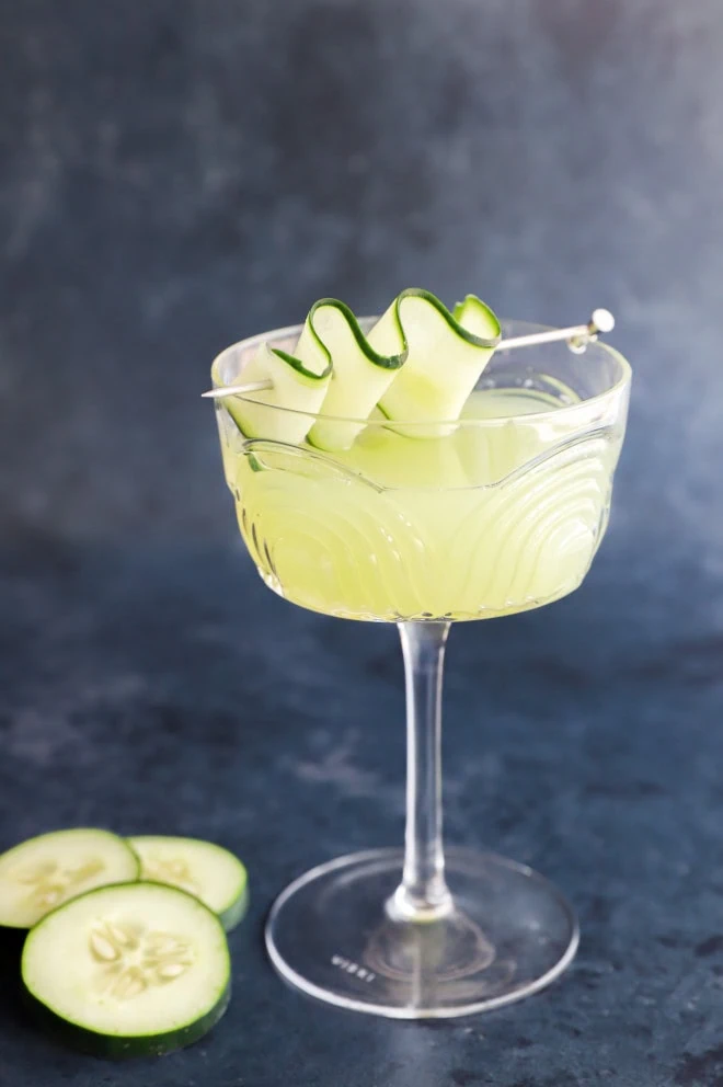 A cucumber martini in a coupe glass with cucumber slices next to it