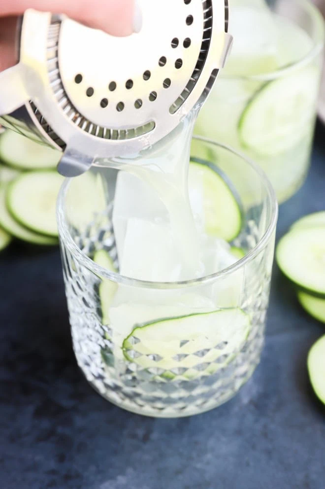 straining cucumber margarita into a cocktail glass with fresh cucumber