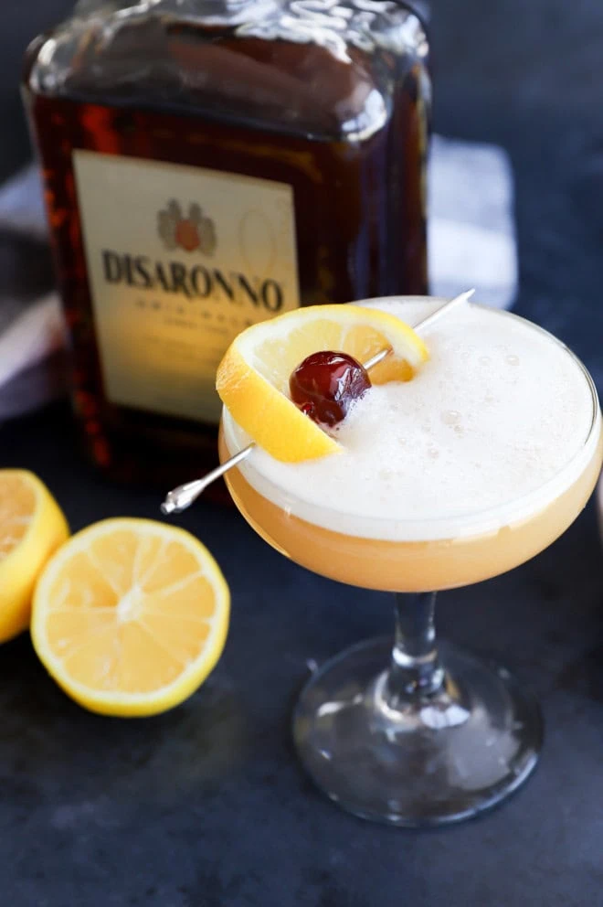 Disaronno sour cocktail in a coupe glass with lemon and cherry garnish and disaronno bottle in the background