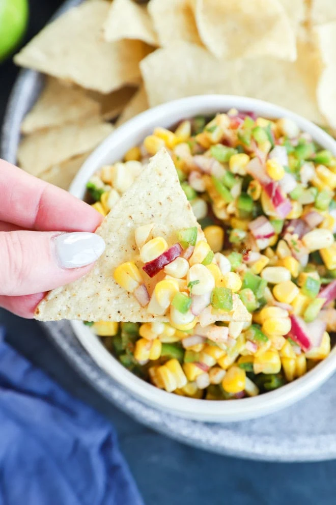 Hand holding a chip with roasted chili corn salsa in a bowl and tortilla chips on a plate