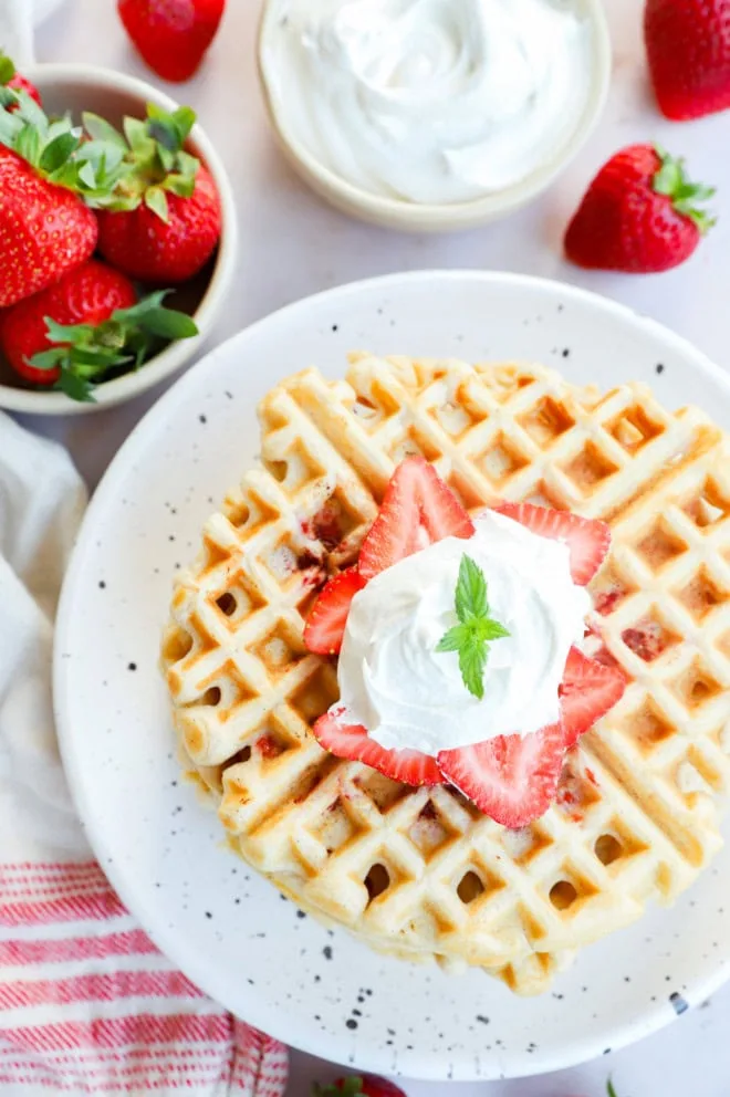 strawberry waffles on a plate with fresh strawberries and whipped cream in bowls