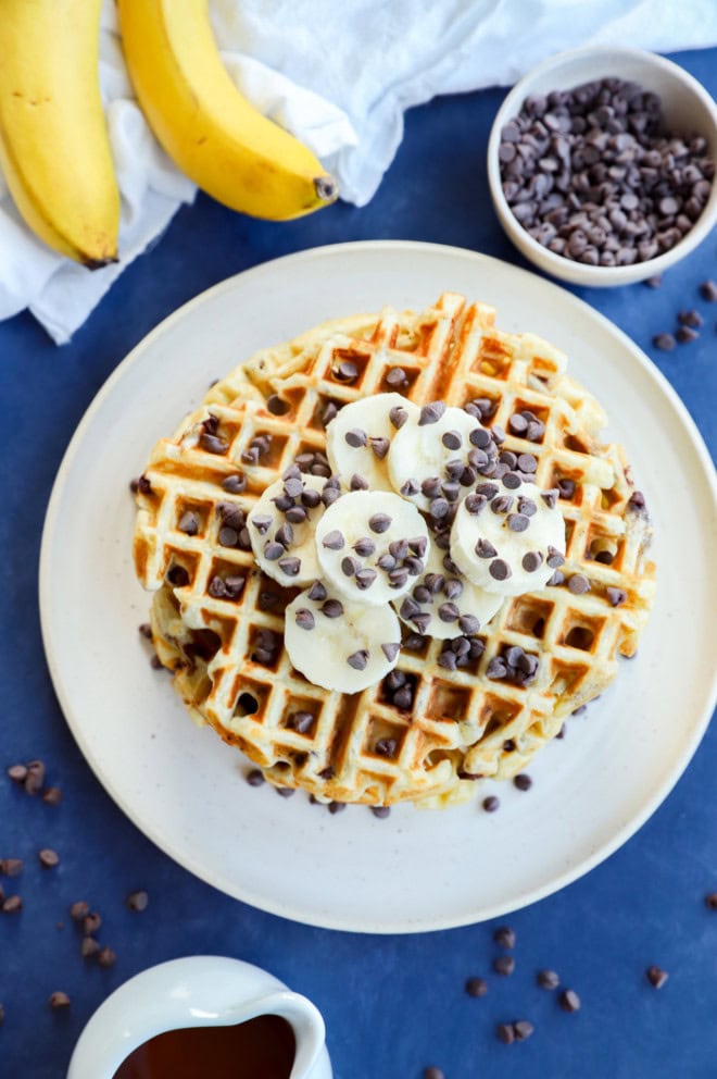 overhead image of banana waffles in a stack on a plate with fresh bananas and a bowl od chocolate chips