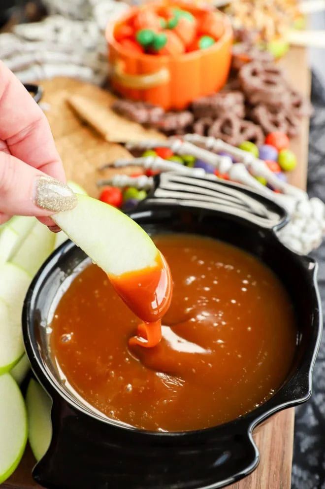 hand holding an apple slice dipping in caramel apple dip with skeleton hand in background