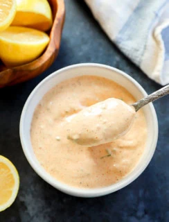 spoon lifting out sauce for crabs cakes from bowl with bowl of lemon halves