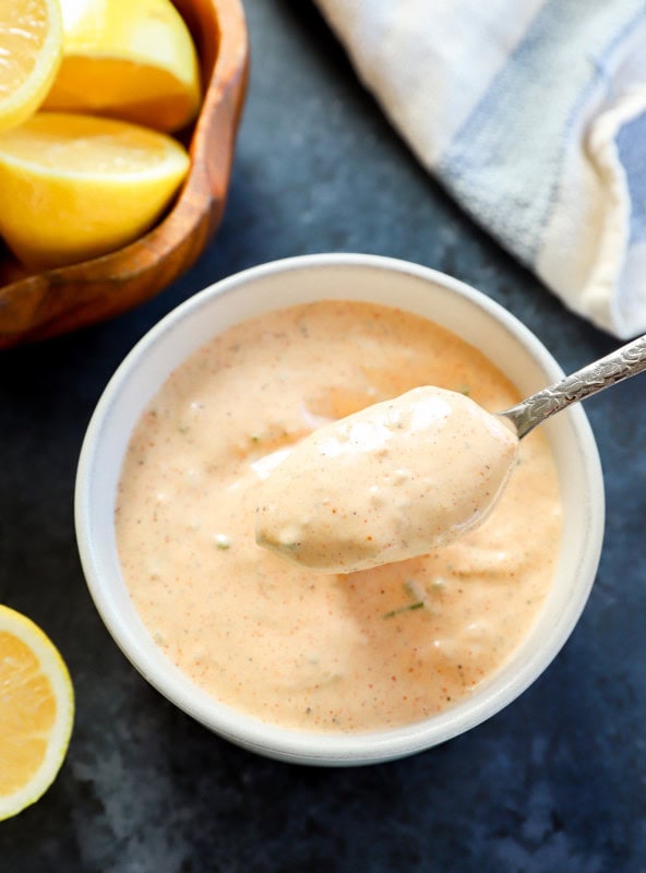 spoon lifting out sauce for crabs cakes from bowl with bowl of lemon halves