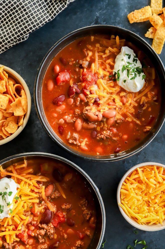 bowls of instant pot chili topped with cheese, sour cream, and parsley with fritos on the side