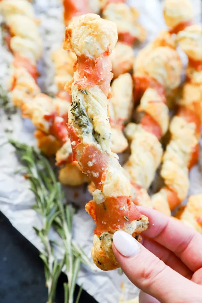 Hand holding prosciutto cheese twists over a pile of them with parchment paper