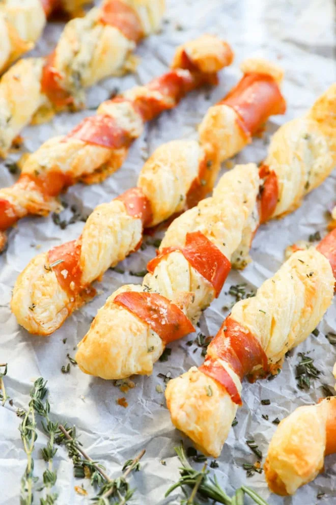 prosciutto cheese twists lined up on a piece of parchment paper with fresh herbs