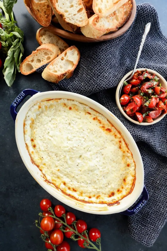 overhead image of hot cheese dip in baking dish with tomato topping in bowl and toasted bread slices