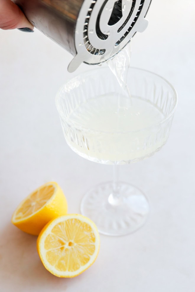 pouring bees knees cocktail into a coupe glass with lemon halves