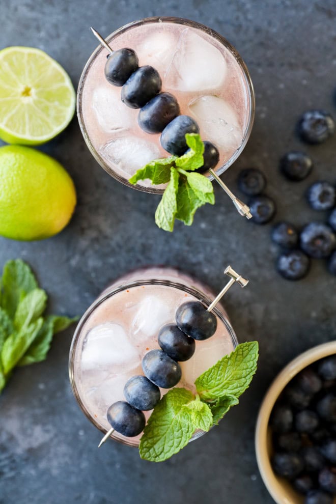 two cocktails with fresh berries and fresh herbs and lime halves